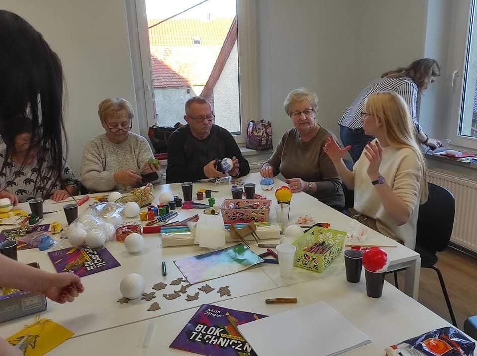 Zdjęcie: Pani Paulina Bagińska wraz z uczestnikami rozmawiająca przy stole, na którym znajdują się artykyuły plastyczne